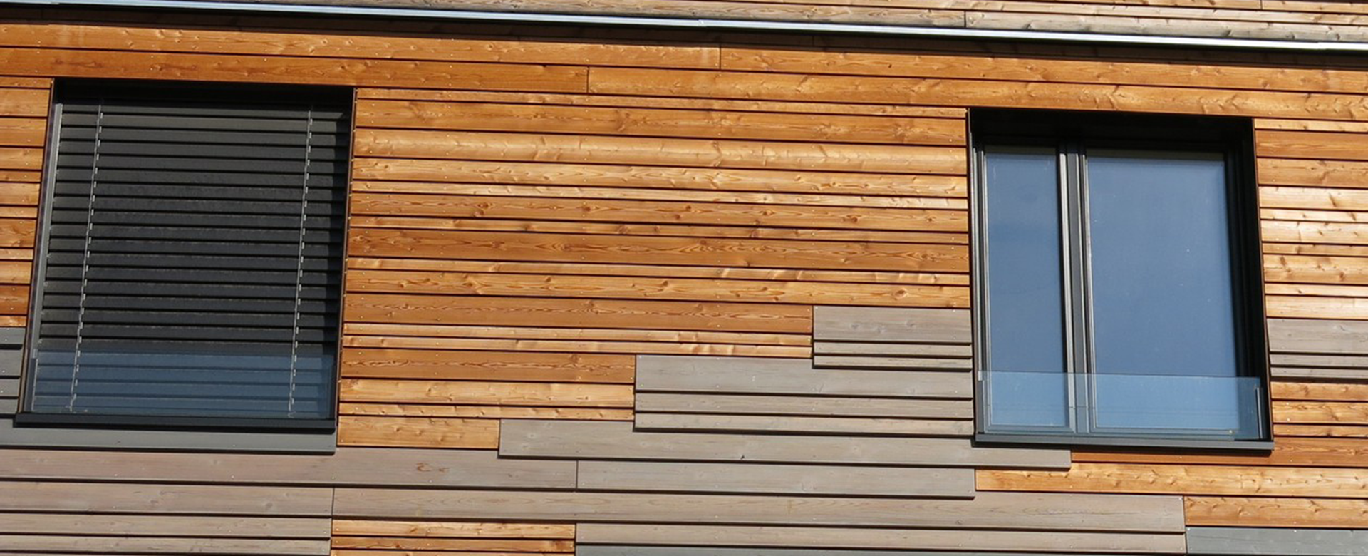 Holzfassade aus unterschiedlichen Holzarten mit zwei Fenstern, eines davon mit Sonnenschtz.